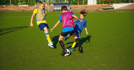 Three young soccer players in colorful training vests compete for the ball on a green field. The action is captured as they make a move, with a goalpost and clear skies in the background.