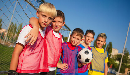 The photo shows five young soccer players posing in front of goal on a sunny day. They wear colorful jerseys and vests, with one player holding soccer ball. Their expressions reflect teamwork and fun