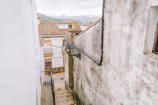 Narrow alley with stone steps, whitewashed walls, rustic rooftops, a streetlamp, and a scenic view of hills in Gaucín, Spain, under a cloudy sky.