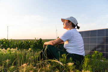 Woman wearing white cap sits next to solar panel at sunset. Wind turbines in the background.