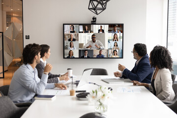 Multiethnic different aged business team speaking to distant colleagues on group conference chat. African American company leader man talking to staff from large display on online and offline meeting