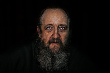 A dramatic close-up portrait of an elderly man with deep wrinkles, a long beard, and piercing eyes. The dark background enhances the intense, emotional expression.