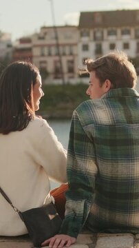 Vertical video, A young couple sits by the Canal de Alfonso XIII in Seville, Spain, enjoying the golden sunlight and each other's company on a warm day.