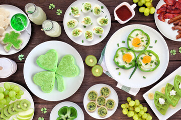 St Patricks Day breakfast or brunch table scene against a dark wood background. Assortment of green and shamrock themed pancakes, toasts, eggs, fruits and meats. Overhead view.