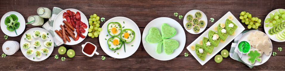 St Patricks Day breakfast or brunch table scene against a dark wood banner background. Assortment of green and shamrock themed pancakes, toasts, eggs, fruits and meats. Top view.