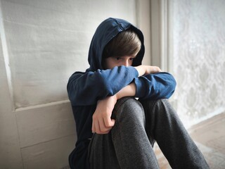 A boy in a sweatshirt sits on the floor in the room. A boy sitting alone with sad feeling.. © Anastasiya
