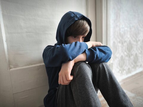 A boy in a sweatshirt sits on the floor in the room. A boy sitting alone with sad feeling..