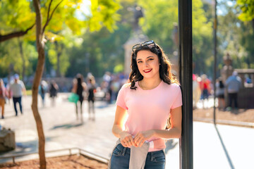 Fototapeta premium Cheerful young Latina woman with wavy hair, pink t-shirt, and jeans leans on a lamppost in Alameda Central, Mexico City, enjoying a sunny day surrounded by nature and people. 