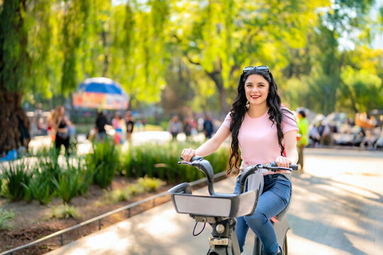 Happy young Latina woman with wavy hair, pink t-shirt, and jeans rides an Ecobici through Alameda Central, Mexico City, enjoying a sunny day surrounded by nature and people.  