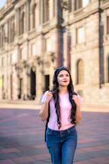 Fototapeta premium Happy young Latina woman with wavy hair, pink t-shirt, jeans, and a red backpack walks in front of the MUNAL museum in Mexico City, enjoying a sunny day and historic architecture. 