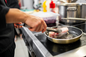 A chef is skillfully using tongs to flip a steak in a frying pan on a modern stovetop. Steam rises, highlighting a busy kitchen environment with various cooking ingredients in the background.