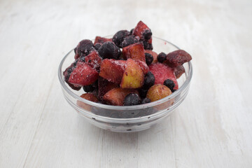 Frozen mixed fruits and berries in a glass bowl on wooden background. Black plum, grapes, apples, mountain ash, grapes.