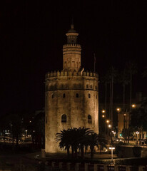 Obraz premium A historic tower stands illuminated at night, overlooking a charming street with outdoor cafés and pedestrians. Warm streetlights add to the cozy, atmospheric cityscape.