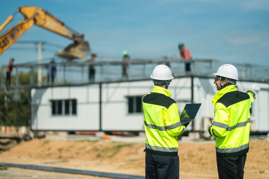 Two engineers wearing high visibility jackets and helmets discussing construction progress at a site, one pointing towards workers and machinery, emphasizing teamwork, safety, and planning in