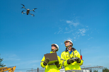 Two engineers in high visibility jackets and helmets observing a drone flying over a construction...