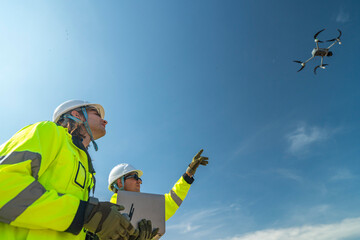Two engineers in high visibility jackets and helmets observing a drone flying over a construction...