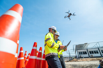 Two engineers in high visibility jackets and helmets operate a drone using a laptop amidst a stack of traffic cones on a construction site. The scene highlights advanced technology and teamwork.