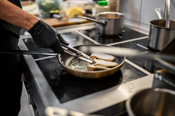 A chef wearing black gloves uses tongs to flip food in a frying pan on a modern stovetop, showcasing a busy kitchen environment with various utensils and pots in the background.