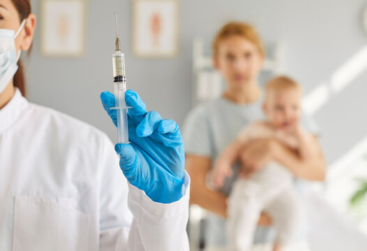 Close up female professional nurse doctor with flu, coronavirus, polio, or hepatitis B vaccine inoculation syringe, mother and infant baby in blurred background. Little children vaccination concept