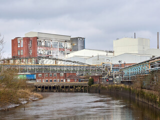 Obraz premium Industrial factory complex with pipes near a river on an overcast day.