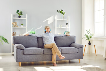 Senior smiling relaxed woman sitting on sofa at home and listening to music or audiobook with headphones. Elderly person wearing casual clothes in earphones resting in the living room.
