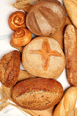 Freshly baked whole grain homemade bread, different types of round sourdough bread with crispy crust, baguettes, buns and ears of wheat on marble background, modern baking concept,