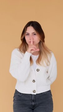 Formal Caucasian young adult woman showing hush gesture with finger on lips on beige background. Concept of a secret. Vertical video.
