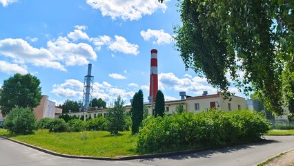 Behind the asphalt path and grassy lawn with bushes, pines and thuja trees growing on it are administrative and industrial buildings with ventilation hoods and factory chimneys. Sunny summer weather