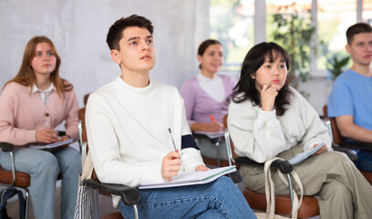 Interested concentrated guy student listening and taking notes of lecture while studying with coursemates in university auditorium