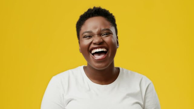 A young African American woman with a joyful expression laughs widely against a bright yellow background in an isolated studio. Her chubby features enhance her cheerful demeanor.