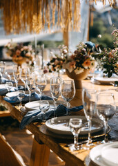 An elegant rustic wedding reception setup featuring a long wooden dining table adorned with refined glassware, white plates, and neatly folded blue napkins.