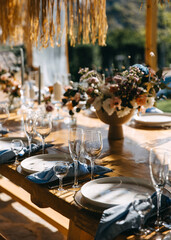 An elegant rustic wedding reception setup featuring a long wooden dining table adorned with refined glassware, white plates, and neatly folded blue napkins.
