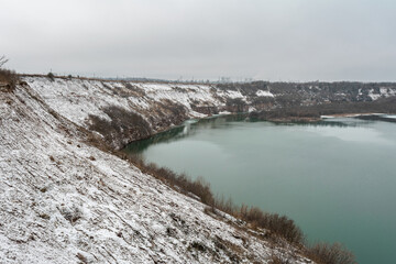 A snow-covered hillside overlooking a serene, blue-green lake. The lake is nestled within a natural crater, surrounded by brown, rocky cliffs. The sky is a pale grey, cold and cloudy winter day.