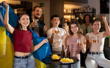 Group of fans of Ukrainian team with a flag are watching the match on TV in a beer bar. They are...