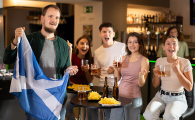 Group of friends fans watching match cheering with Scottish flag in bar