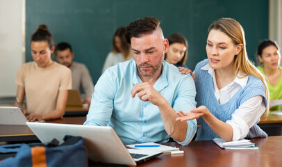 Young student studying with a girl fellow student in a university auditorium is looking for information on a laptop, ..discussing current issues of study