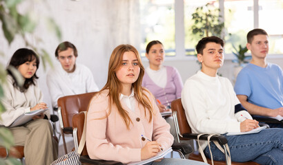 Fototapeta premium Interested motivated girl student listening with attention and taking notes of lecture while studying with coursemates in university auditorium