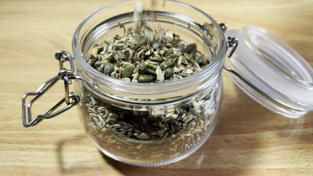 Closeup slow motion shot of mixed edible seeds (sunflower, pumpkin, hemp and linseed) pouring into an open glass storage jar, with an airtight lid and metal clasp. 