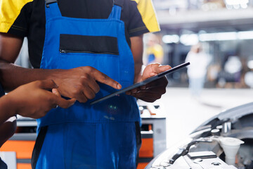 African american engineer trainee uses tablet in car service to watch tutorial on how to replace motor. Garage workspace intern looks up training videos online on device, unsure how to fix vehicle