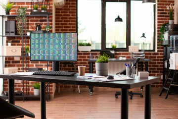 Computer screen displaying financial documents sits on spacious desk in startup workspace. Desktop pc on office table with screen showing business analytics in vacant brick wall room.
