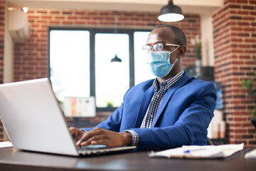African american manager with face mask, typing and replying to work emails on his laptop in brick...