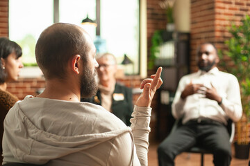 Closeup of bearded man making different signs with fingers during group therapy session. Young white guy sitting in circle and doing hand gestures in brick wall room as people discuss in background.
