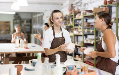 Pleased young man and woman in pinafore working with ceramic dish using tool in workroom