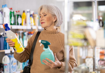 Uncertain mature female buyer choosing convenient spray surface cleaner in department store