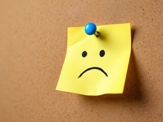 A sad face drawn on a yellow sticky note pinned to a corkboard, conveying disappointment.
