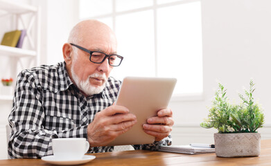 Senior man reading news on digital tablet. Serious mature male using portable computer at home, sitting at table, copy space