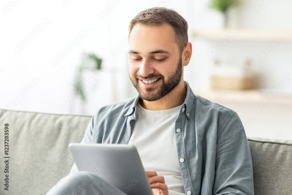 Poster joyful millennial man sitting on sofa with tablet computer, working or studying online at home. happ