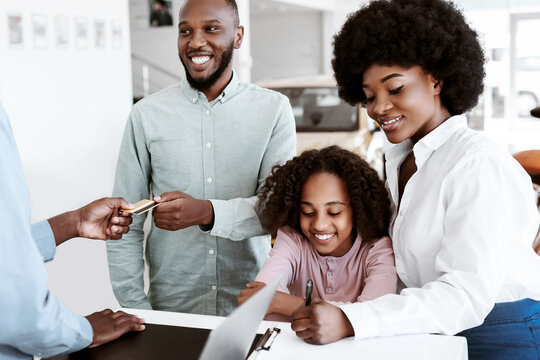 Joyful black family giving credit card to car salesman, paying for new auto at dealership store. African American customers purchasing automobile, signing agreement at showroom