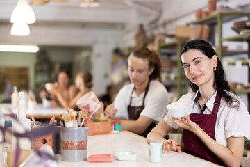 Young woman sits near table and decorates clay ceramic craft vase, plate, works in company of like-minded people. Hand made art, hobbies and creativity.