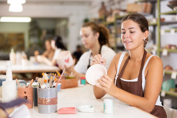 Young woman sits near table and decorates clay ceramic craft vase, plate, works in company of like-minded people. Hand made art, hobbies and creativity.